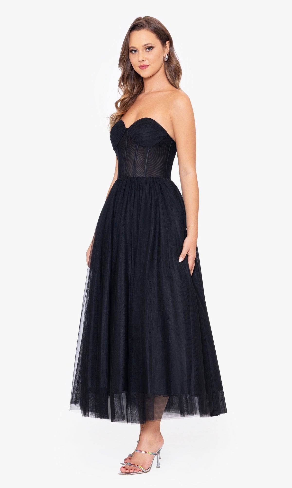 A woman stands against a plain white background wearing the Black Strapless Tea Length Wedding-Guest Dress with a corset bodice and silver high-heeled sandals—an elegant choice for any wedding guest.